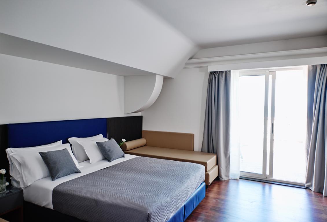 Camera moderna con letto matrimoniale e divano, luce naturale dalle finestre.