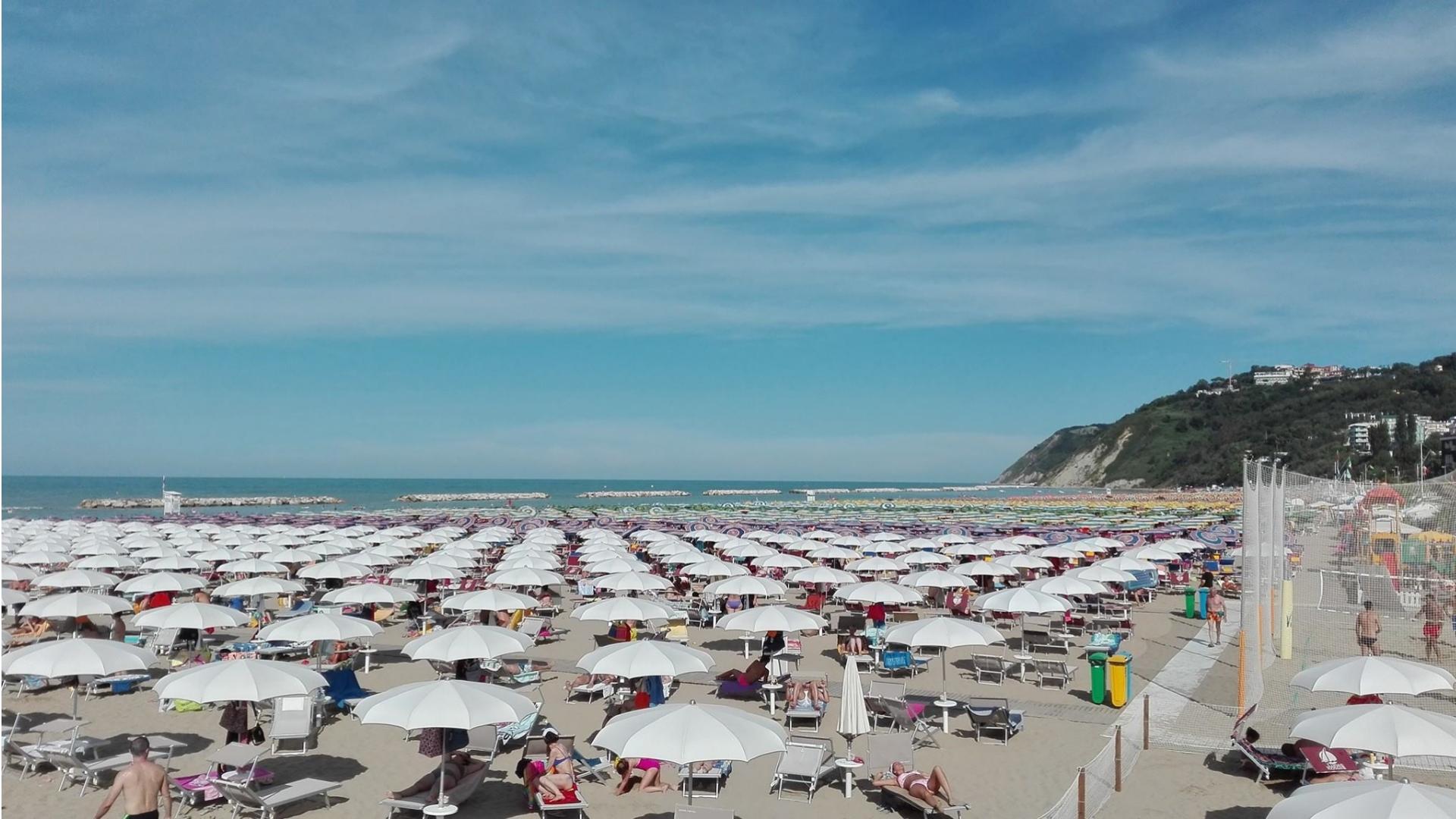 Spiaggia affollata con ombrelloni bianchi e mare calmo.
